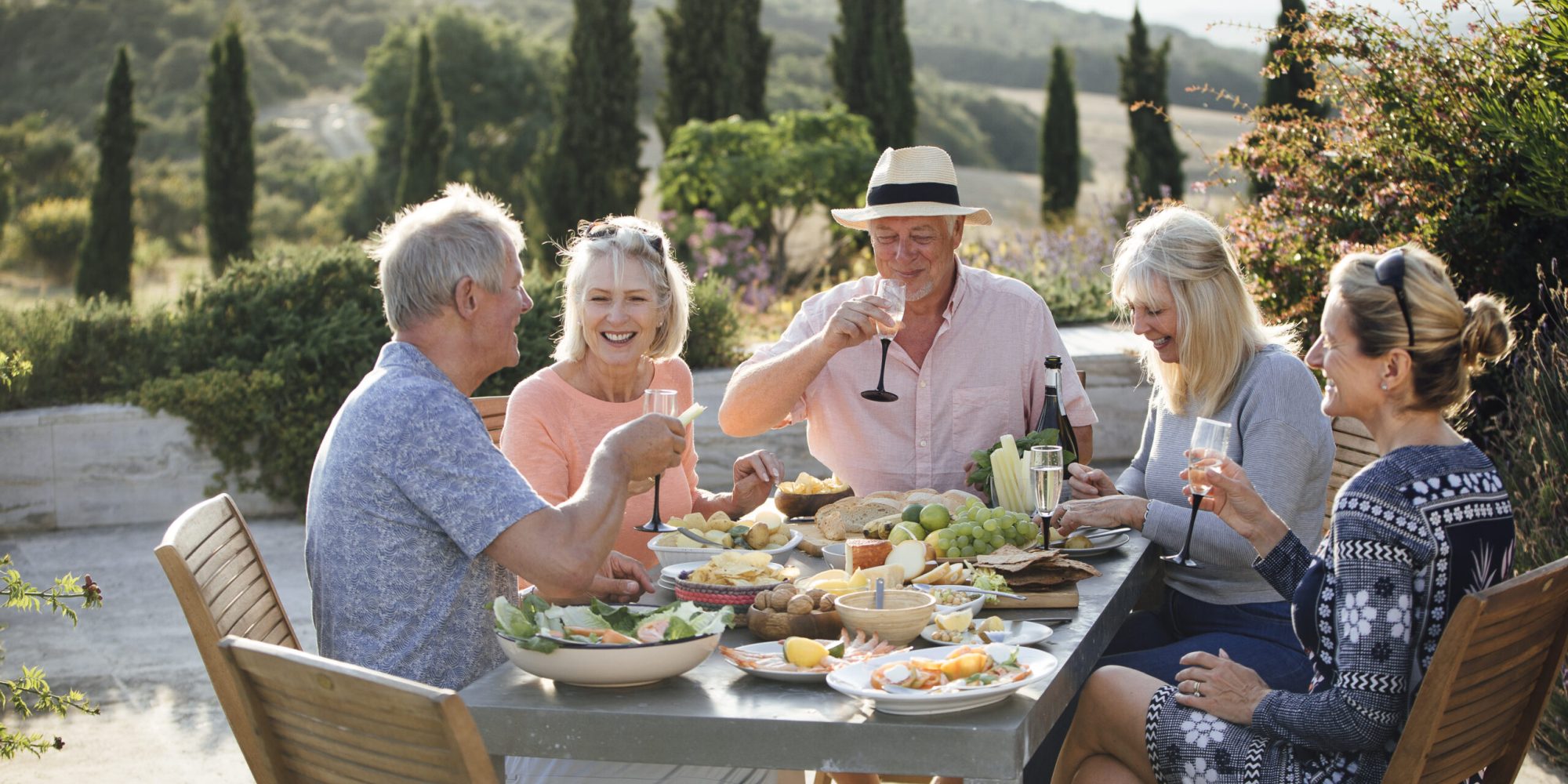 A group of mature friends are sitting around an outdoor dining table, eating and drinking. They are celebrating their holiday with a glass of champagne and enjoying each others company. The image has been taken in Tuscany, Italy.